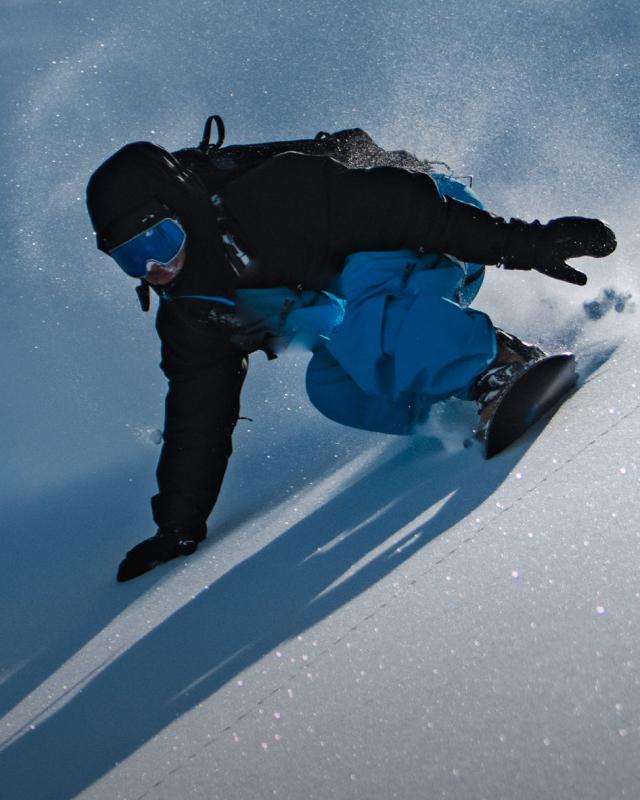 A snowboarder in a black jacket and blue pants carves a turn, kicking up snow.