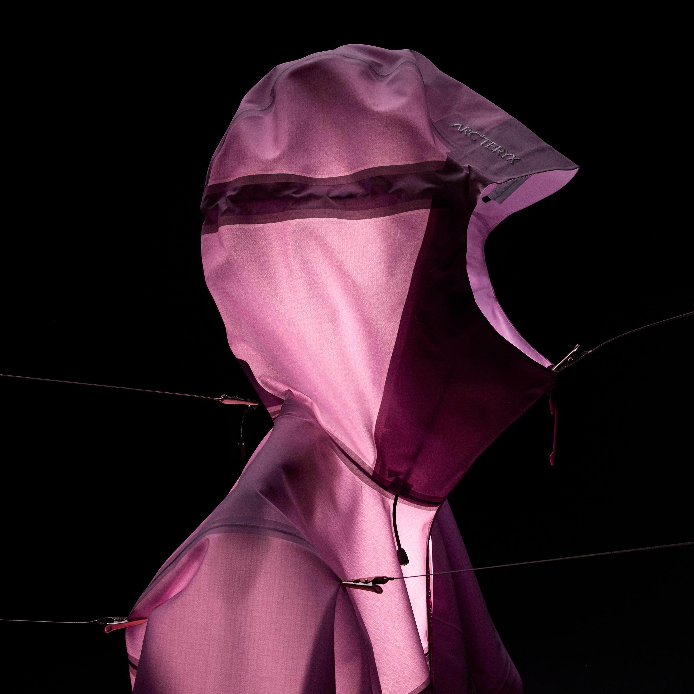 Backlit pink Arc'teryx hooded jacket hanging on wires against a black background.