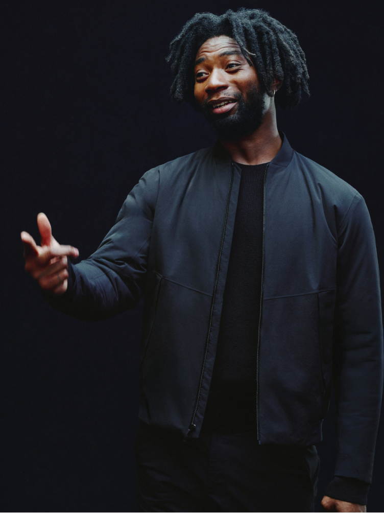 A Black man with dreadlocks and a beard, wearing a dark bomber jacket, smiles and gestures with his left hand against a dark background.