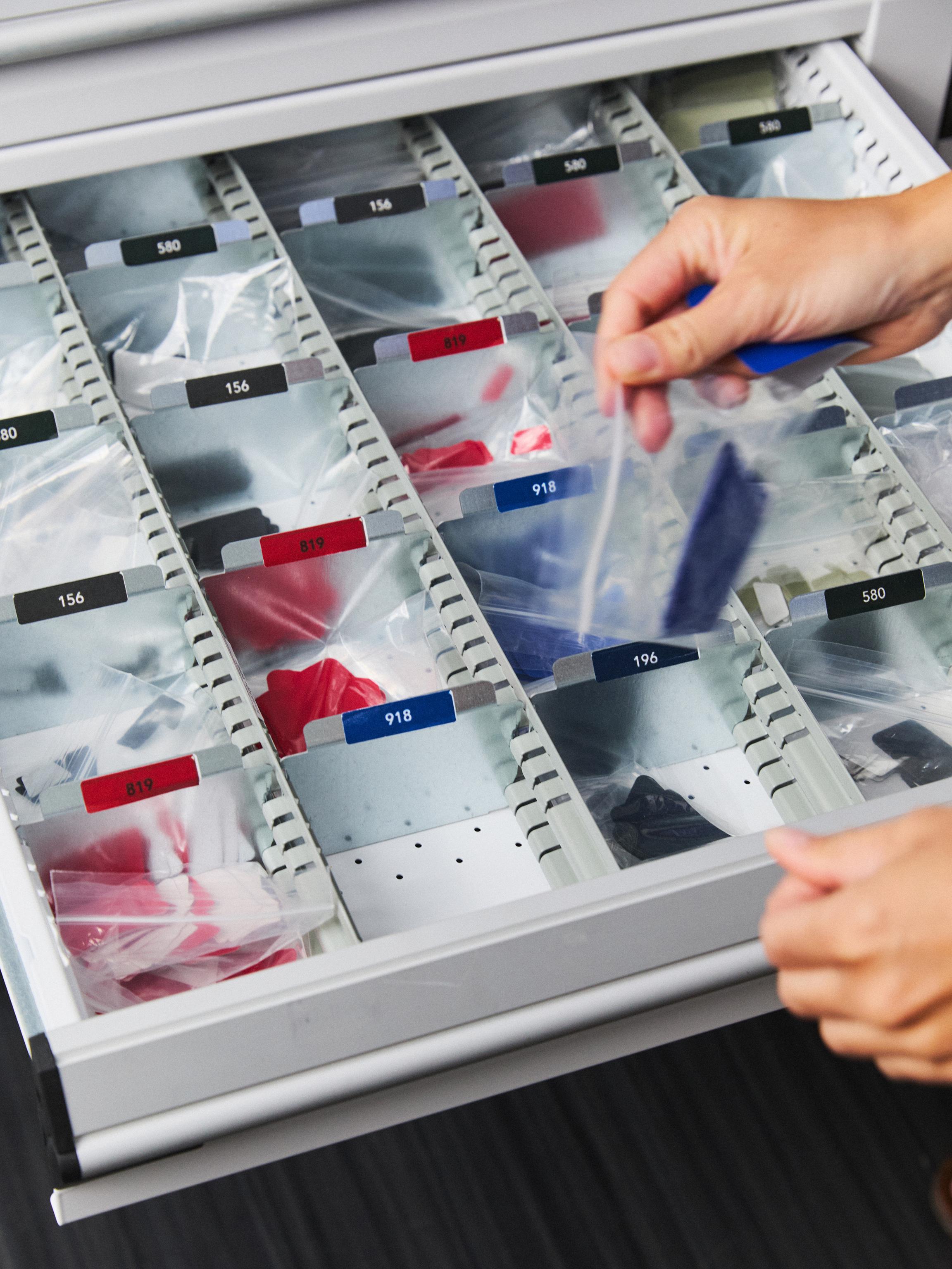 A hand pulls a dark blue fabric swatch in a clear plastic bag from a drawer of organized colorful samples.