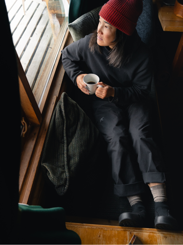 A woman in a red beanie and black outfit holds a coffee mug while looking out a window.