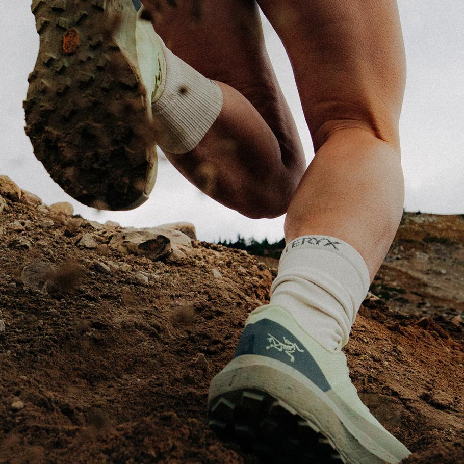 Person's legs and muddy Arc'teryx trail shoes running on a dirt path.