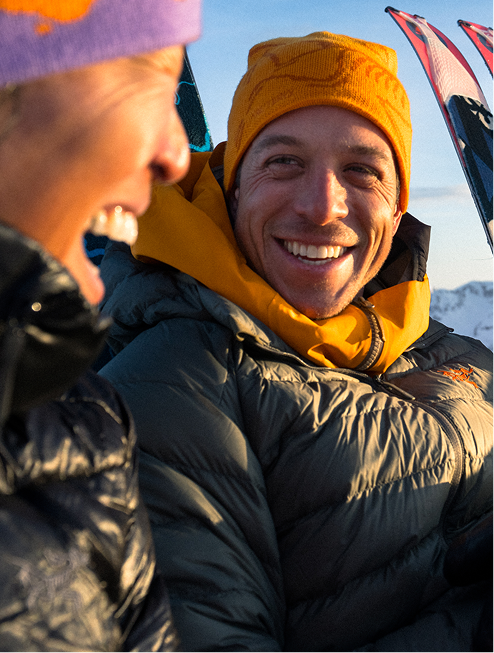 A smiling man in a yellow beanie and dark green puffer jacket looks at a partially visible person with a purple beanie, with skis in the background.