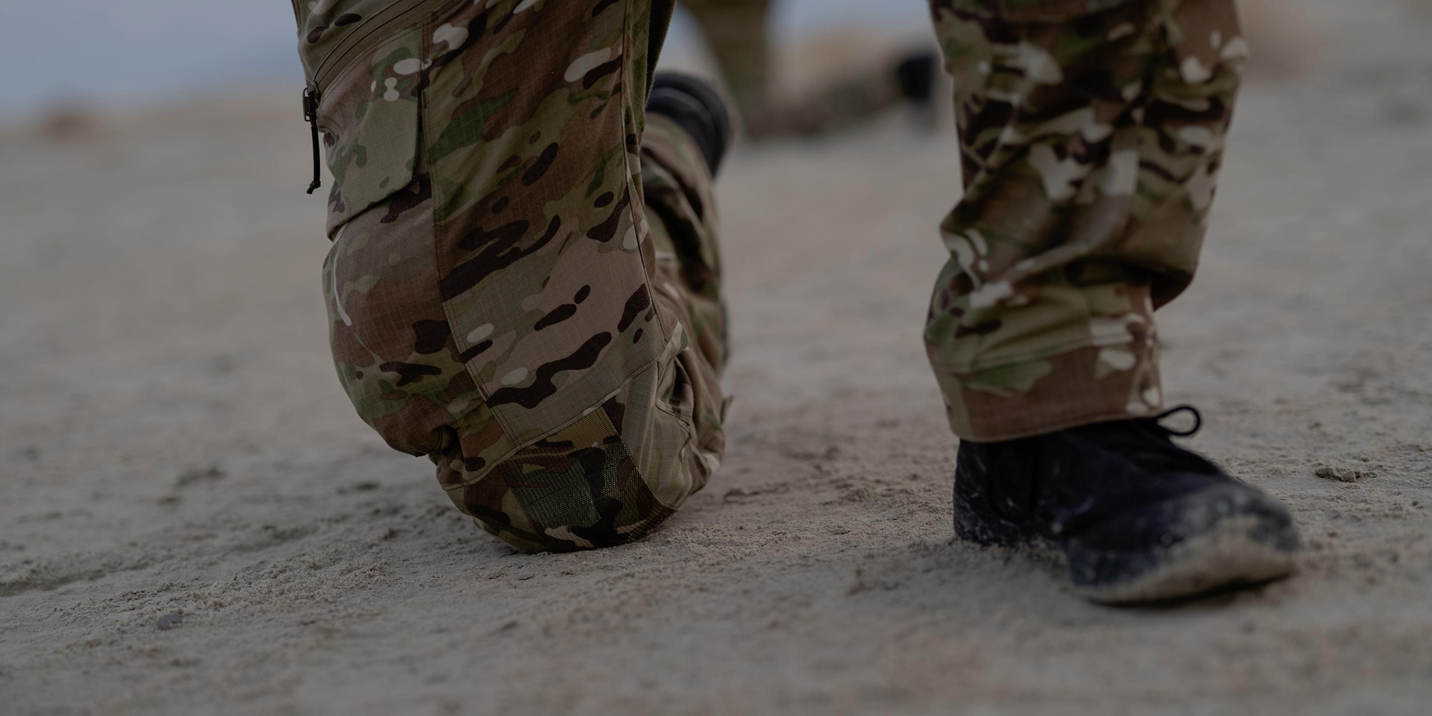 A person in camouflage pants kneels on sandy ground.