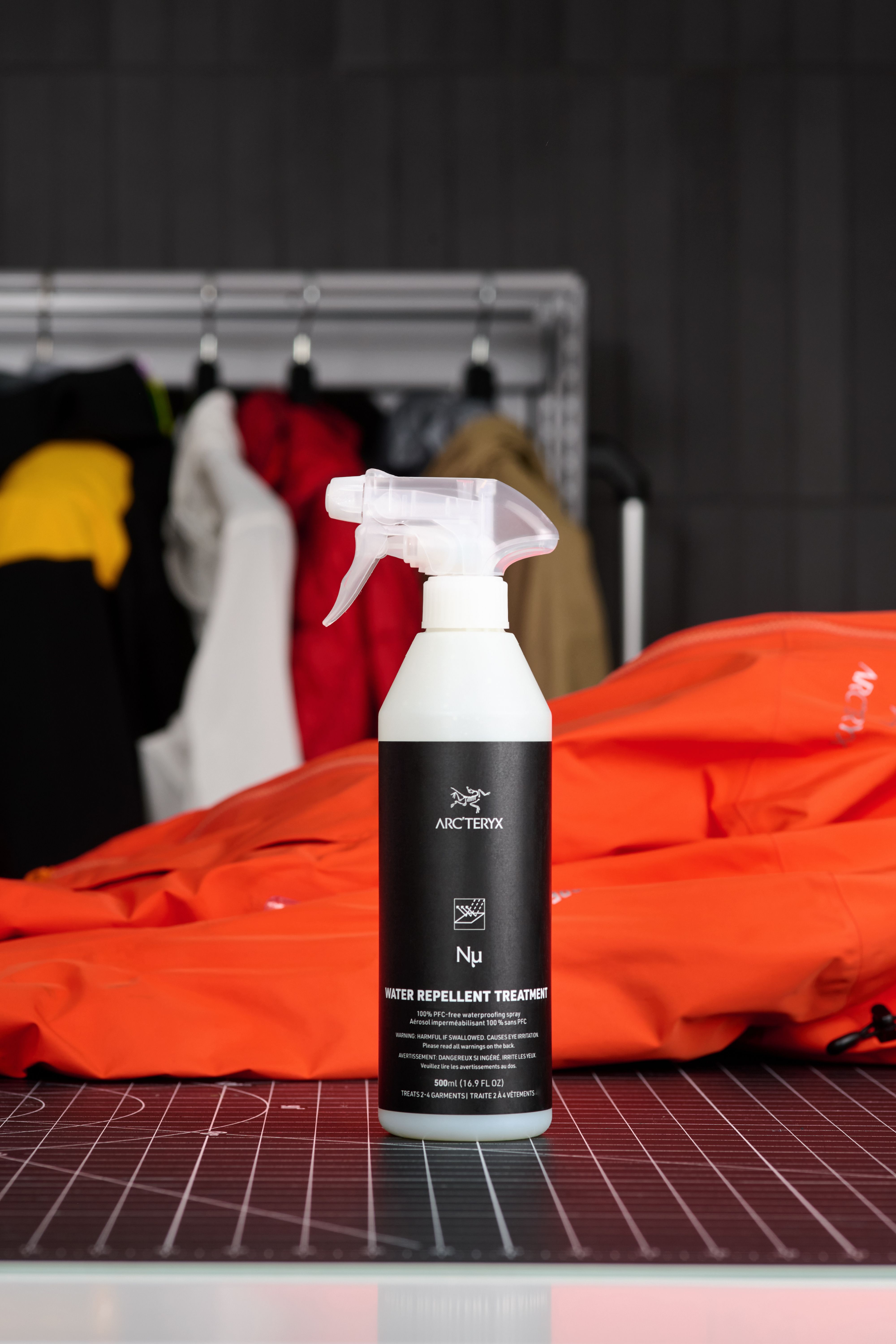 Arc'teryx Nμ Water Repellent Treatment spray bottle next to a folded orange jacket, with other jackets blurred in the background.