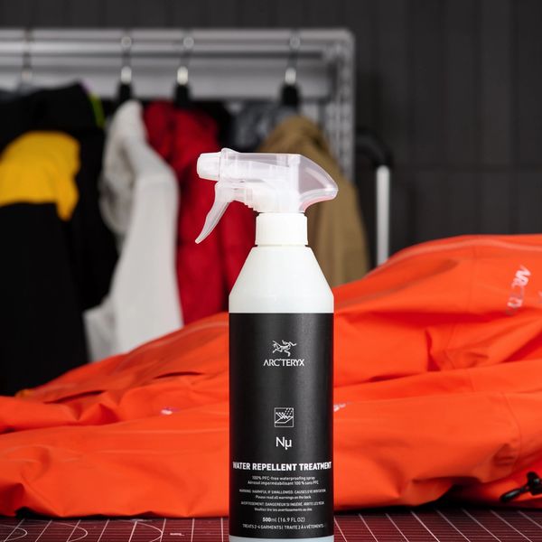 A white spray bottle of Arc'teryx Nµ Water Repellent Treatment sits on a black grid mat, with an orange jacket and blurred colorful jackets in the background.