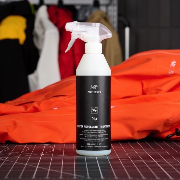 Arc'teryx Nμ Water Repellent Treatment spray bottle next to a folded orange jacket, with other jackets blurred in the background.