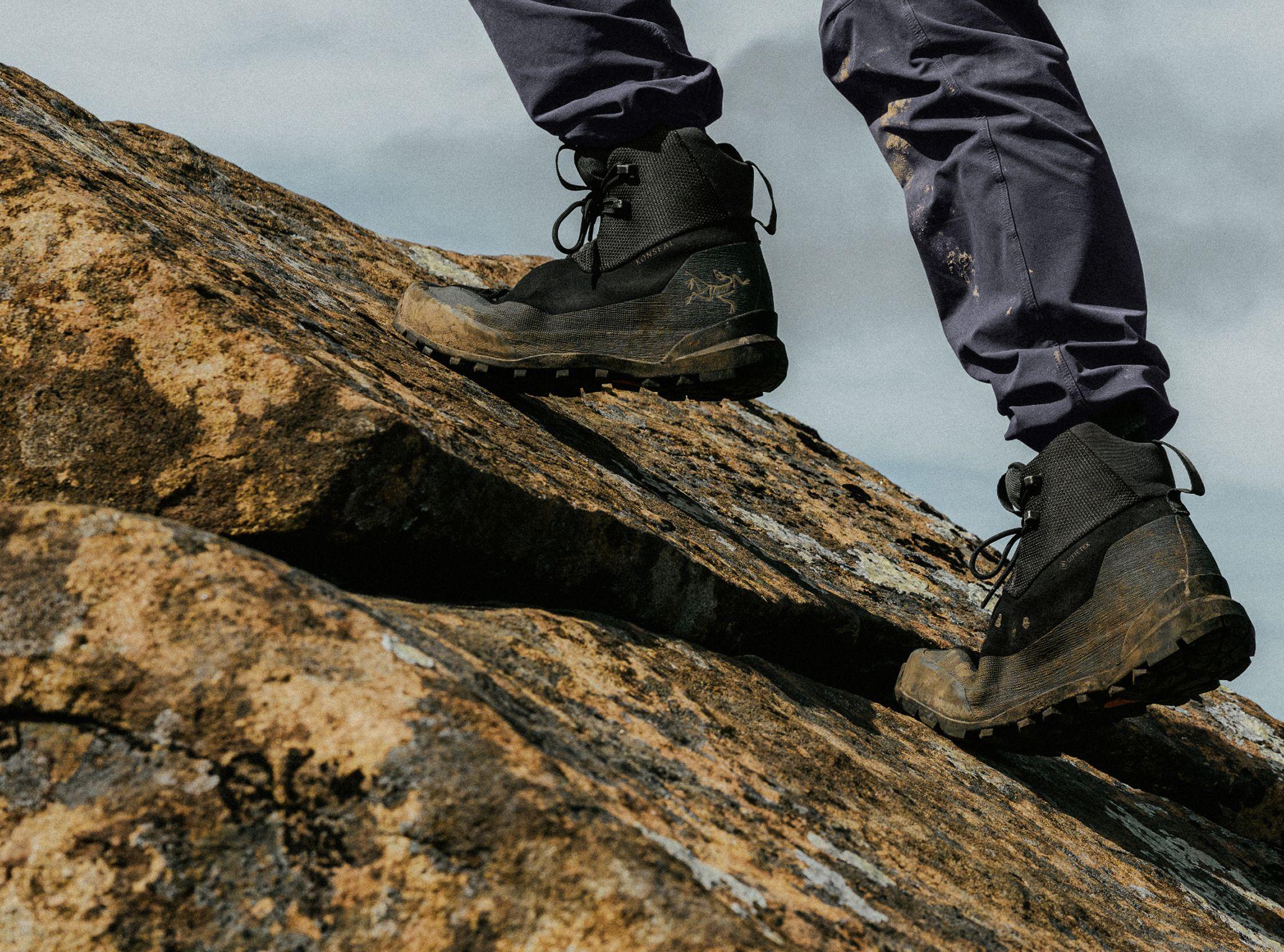 A person's legs and feet in dirty dark hiking boots and pants climbing a rocky slope.