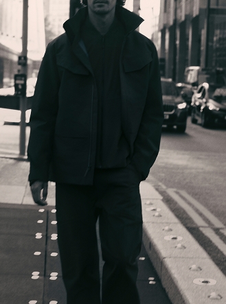 A man in a dark jacket and trousers walks on a city sidewalk, hand in pocket, with buildings and cars in the background.