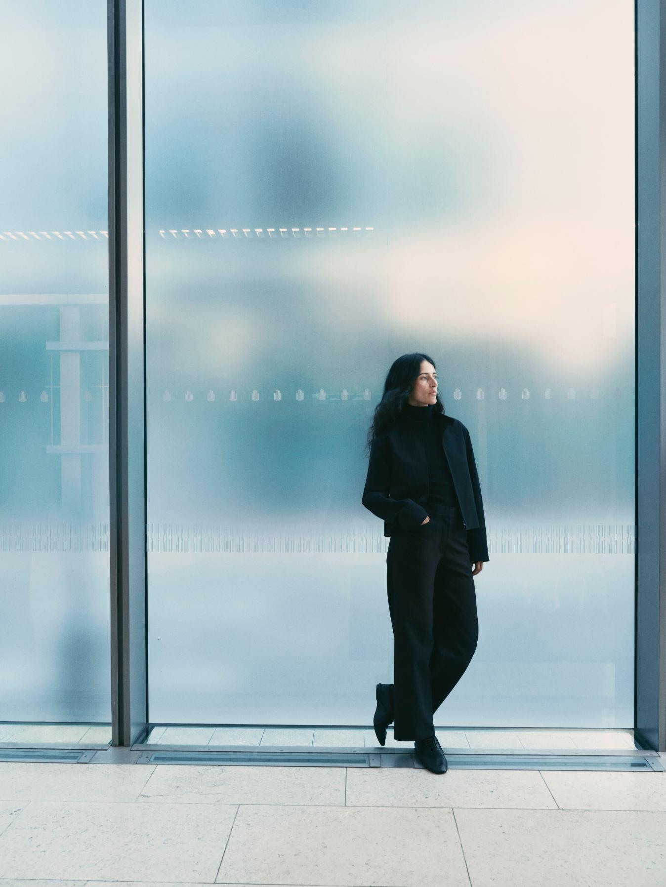 A woman in black clothing leans against a frosted window, looking to the right.