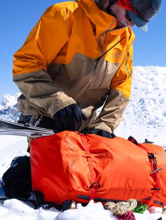 A person in a winter jacket and gloves adjusts an orange gear bag in the snow, with skis nearby.