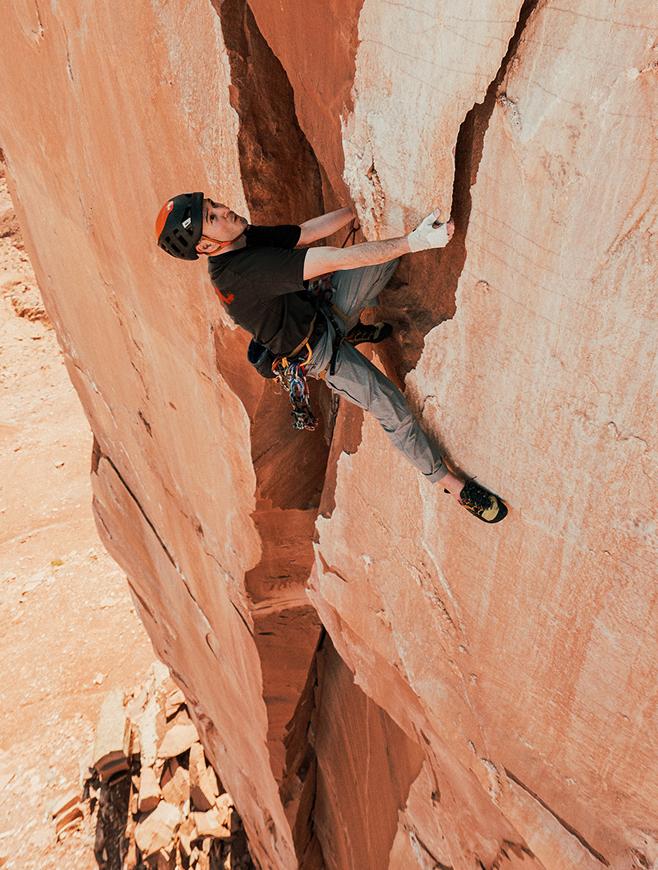 A man in a helmet and climbing gear ascends a reddish-orange rock face, gripping a crack.