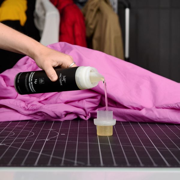 A hand pours Arc'teryx Nyu Technical Detergent into a measuring cup, next to a pink jacket.