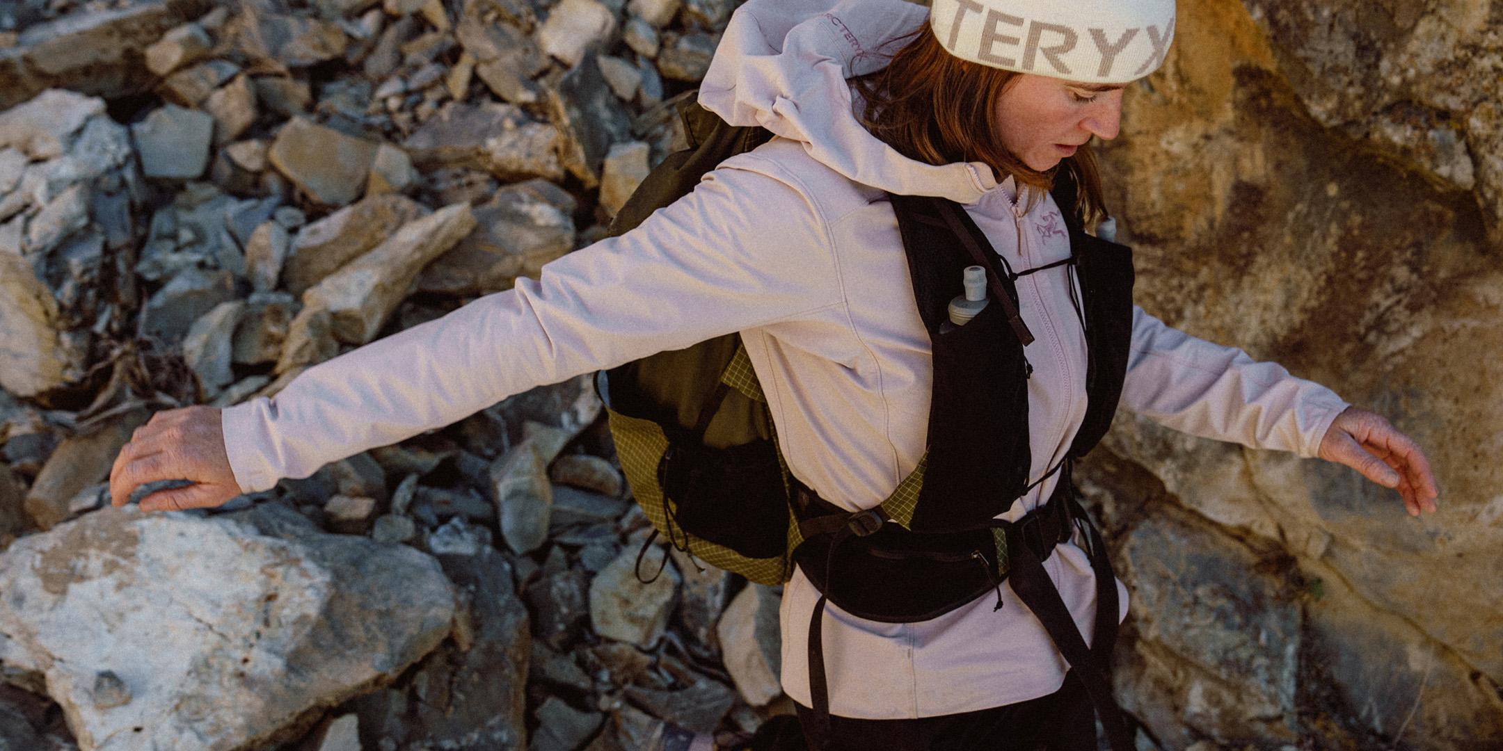 A woman in a light jacket, beanie, and hydration vest carefully walks on rocky terrain.