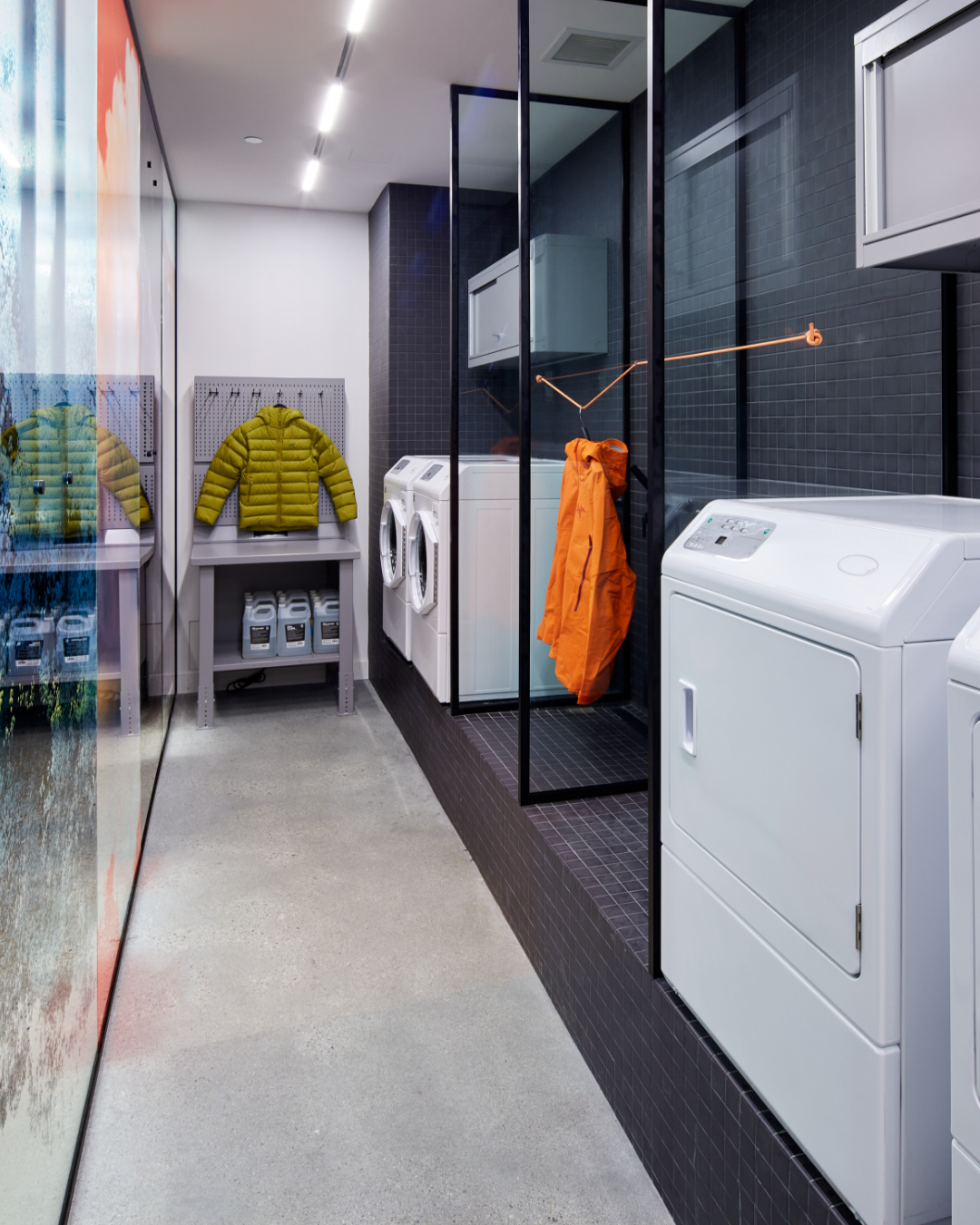 A modern laundry room with white washers and dryers, a yellow puffer jacket on display, and an orange jacket hanging on a glass partition.
