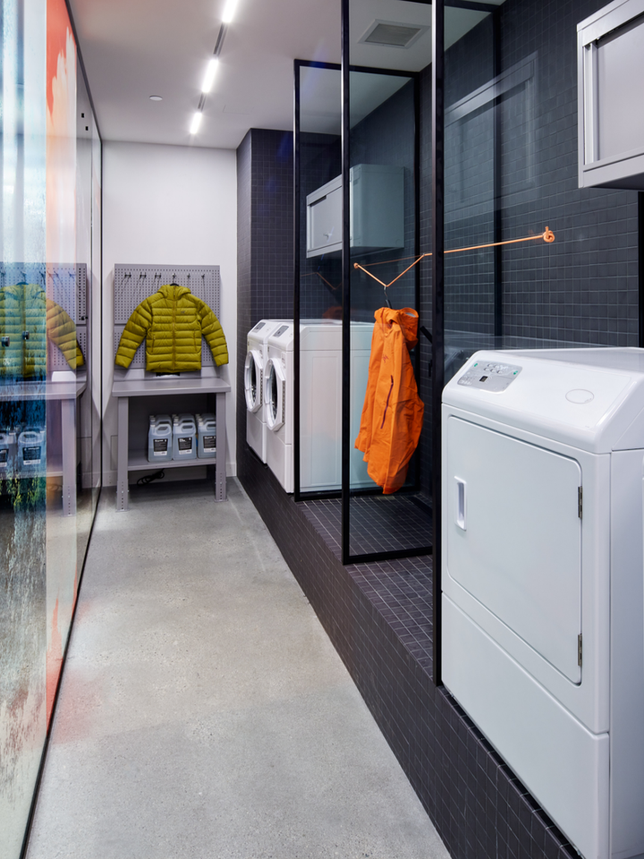 A modern laundry room with white washers and dryers, a yellow puffer jacket on display, and an orange jacket hanging on a glass partition.