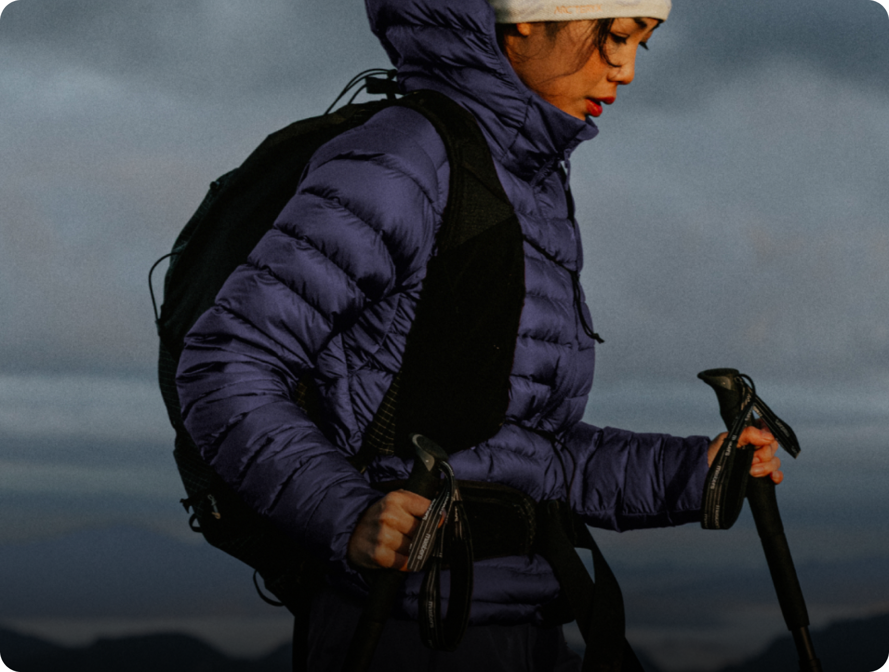 A woman hiking in a purple puffer jacket, white beanie, backpack, and trekking poles.