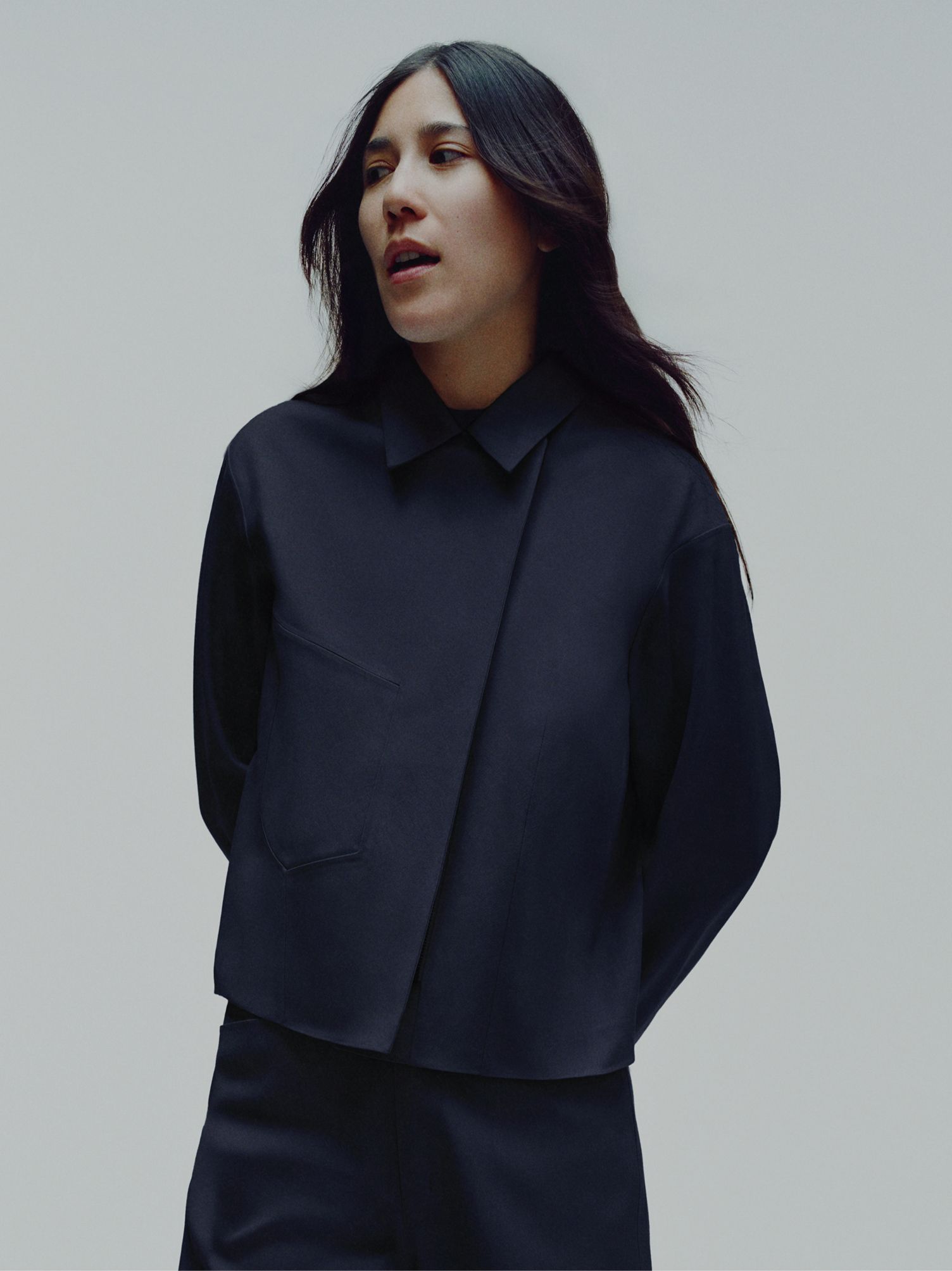 A woman with long dark hair wearing a dark blue asymmetric collared shirt with a chest pocket, looking slightly left.