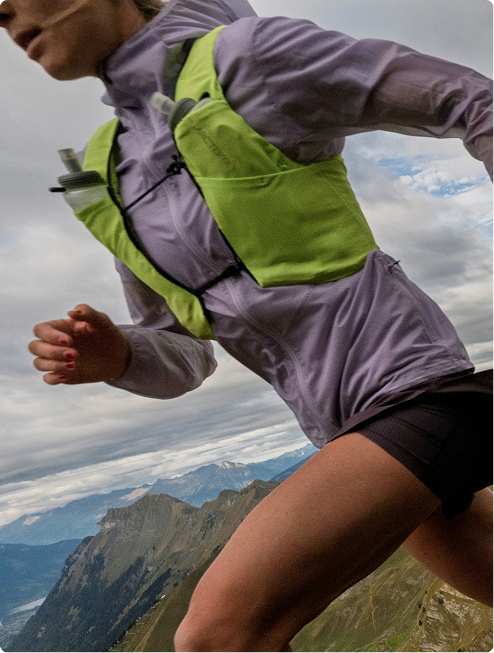 A runner in a purple jacket and green Arc'teryx hydration vest with water bottles, on a mountain trail under a cloudy sky.