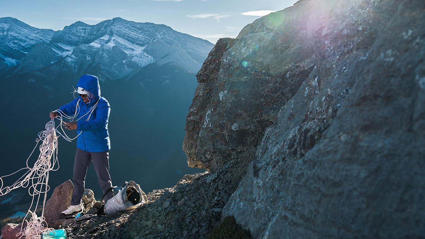 a person in a blue insulated jacket is standing on top of a mountain unraveling climbing rope