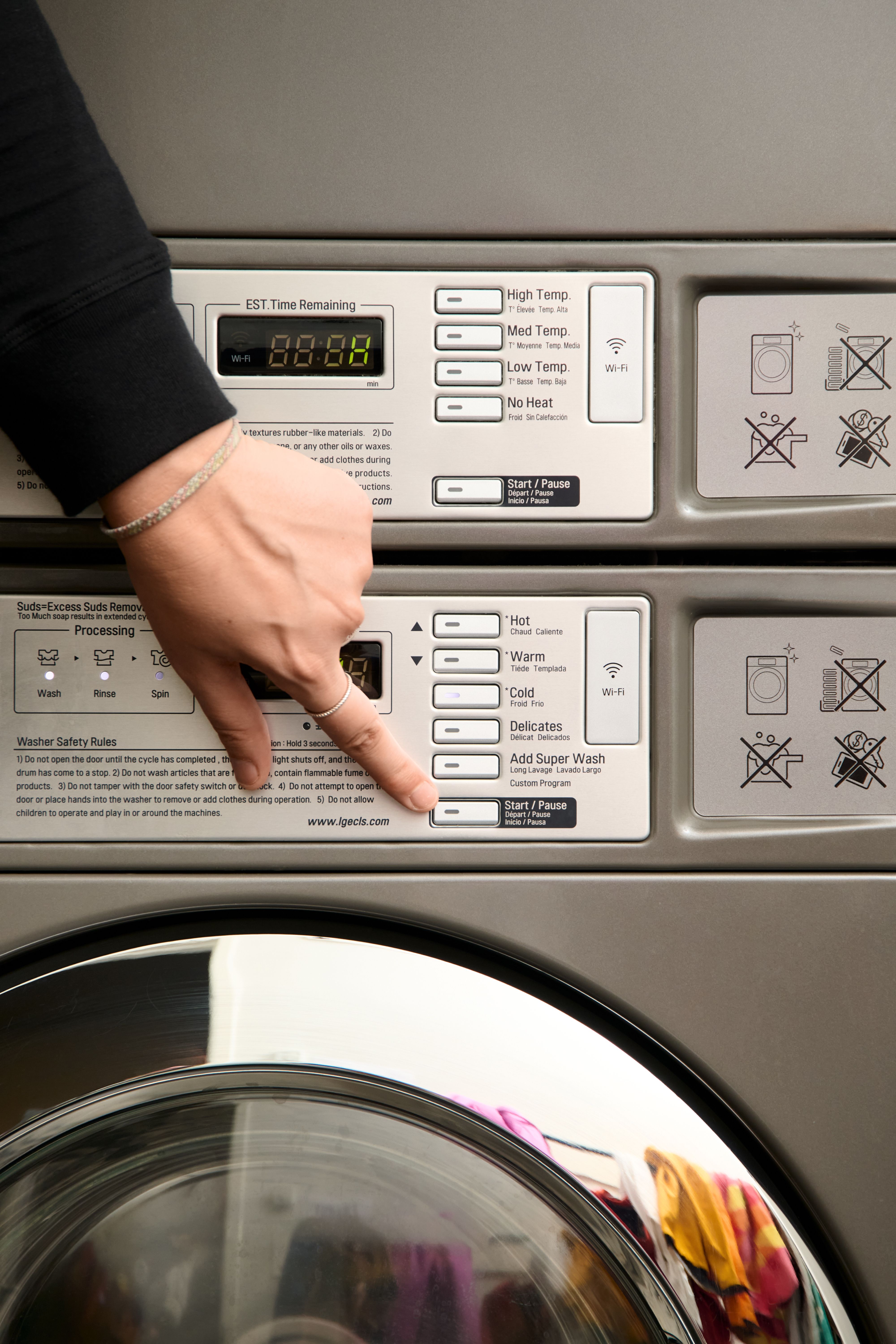 A person's hand presses the Start/Pause button on a washing machine, with colorful laundry visible through the door.