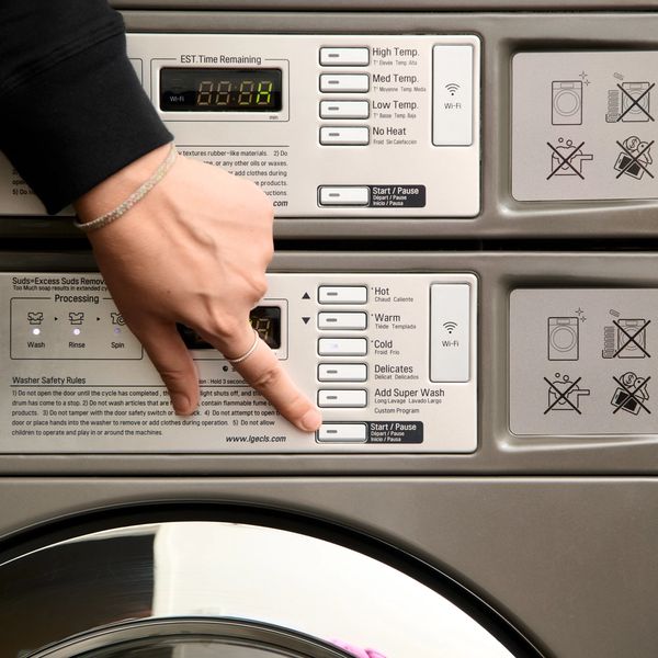 A hand presses the Start/Pause button on a front-loading washing machine's control panel, with colorful clothes visible through the door.