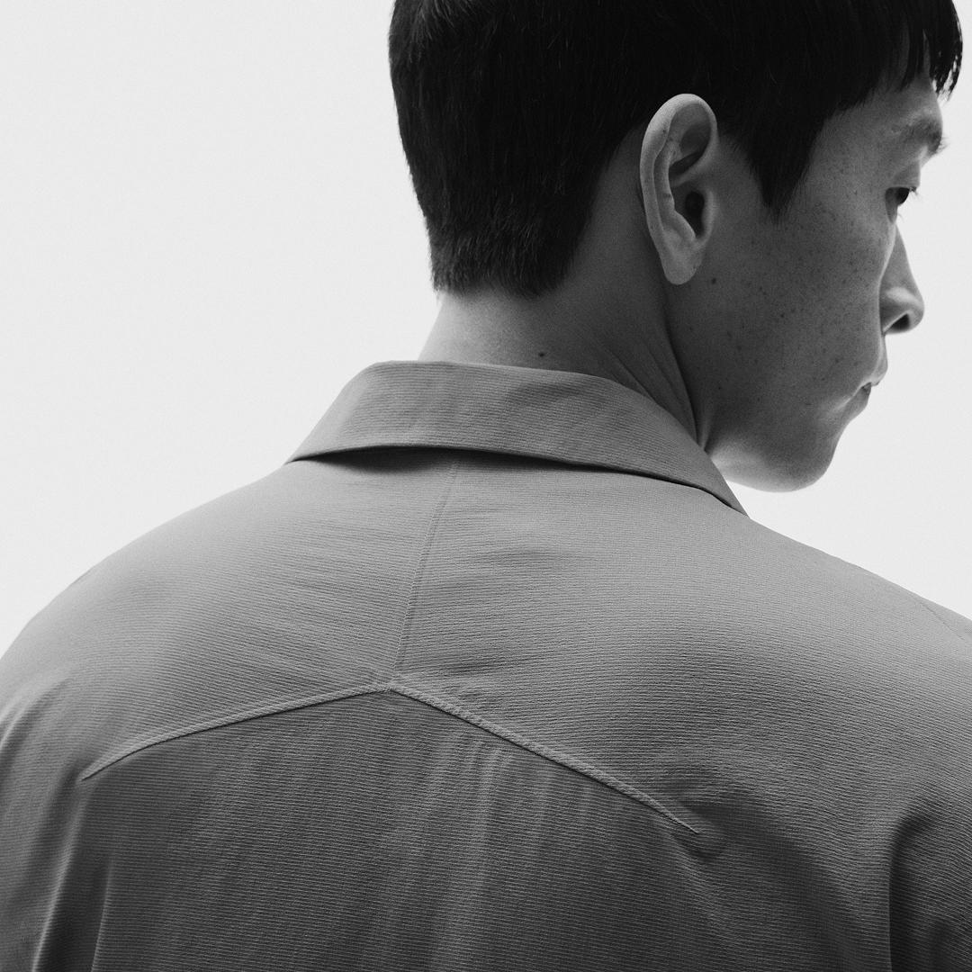 Black and white photo of a man seen from behind, head turned in profile, wearing a collared shirt with a V-shaped seam on the upper back.
