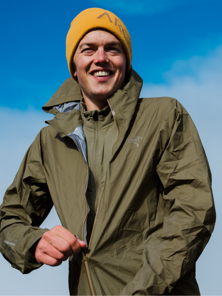 Smiling man in an Arc'teryx jacket and beanie zipping up his coat against a blue sky.