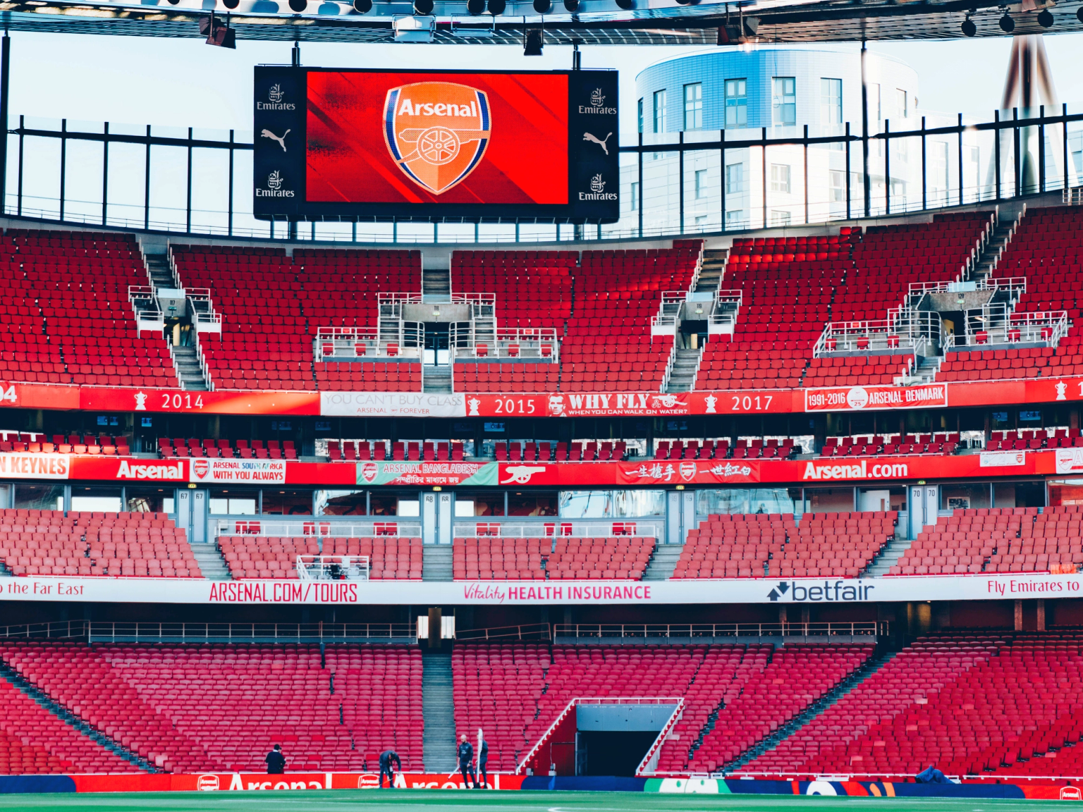 An empty Arsenal football stadium with red seats, a green pitch, and a large screen displaying the Arsenal logo.