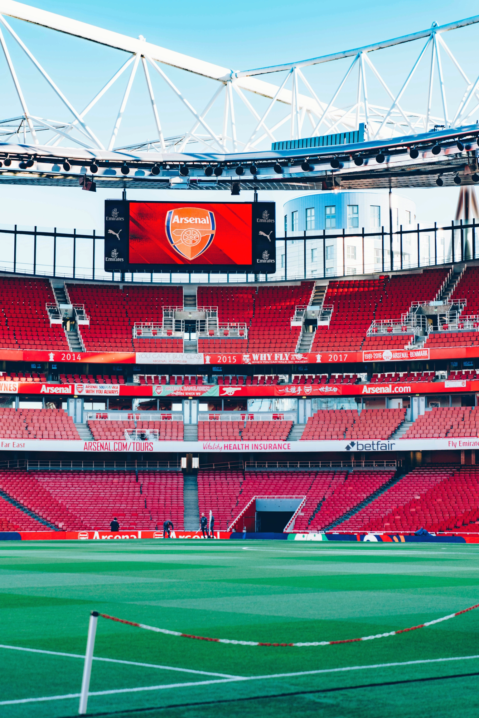 An empty Arsenal football stadium with red seats, a green pitch, and a large screen displaying the Arsenal logo.