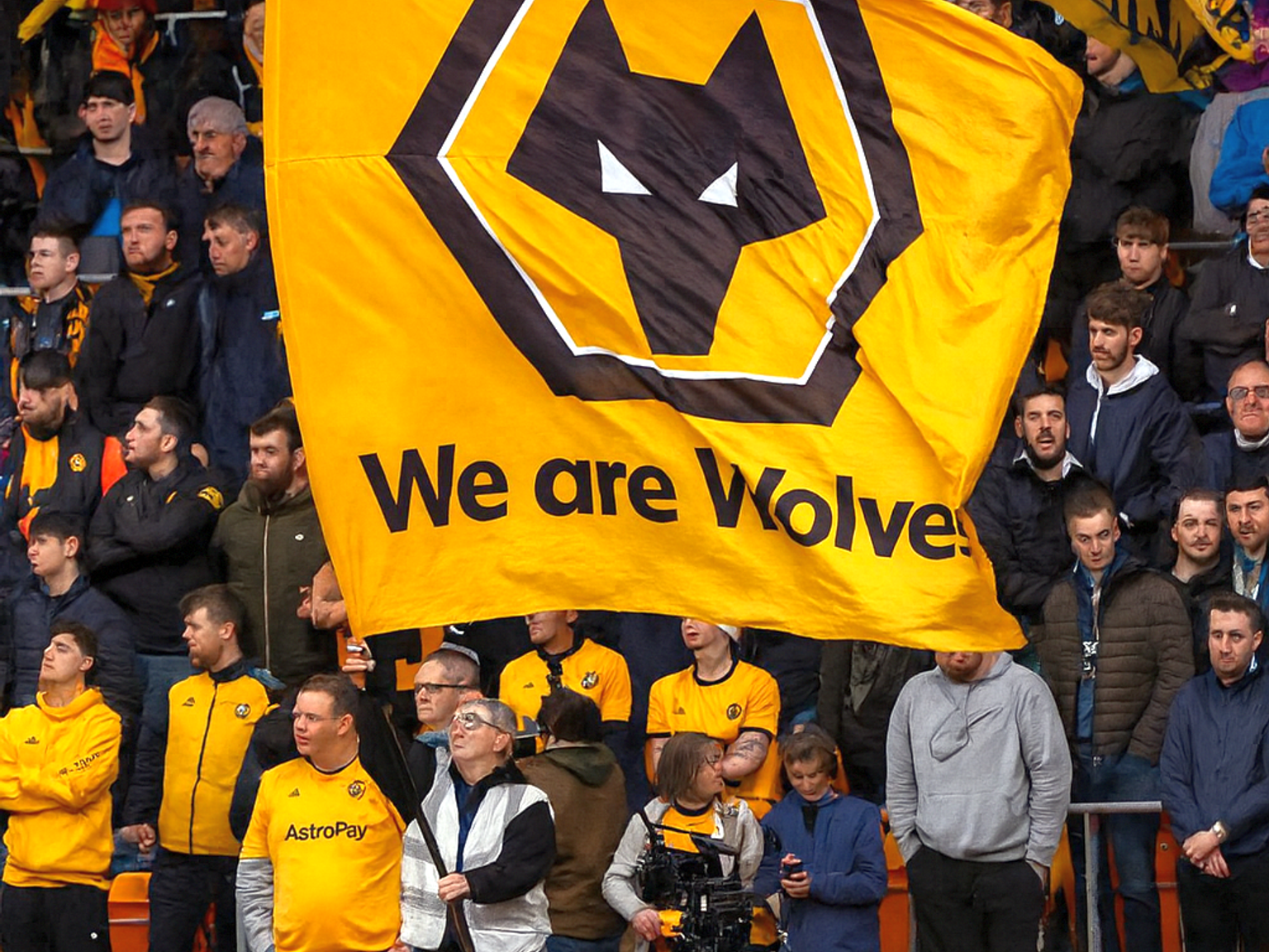 Une foule de fans de football dans un stade, avec un grand drapeau jaune des Wolverhampton Wanderers portant l'inscription 'We are Wolves' au premier plan.