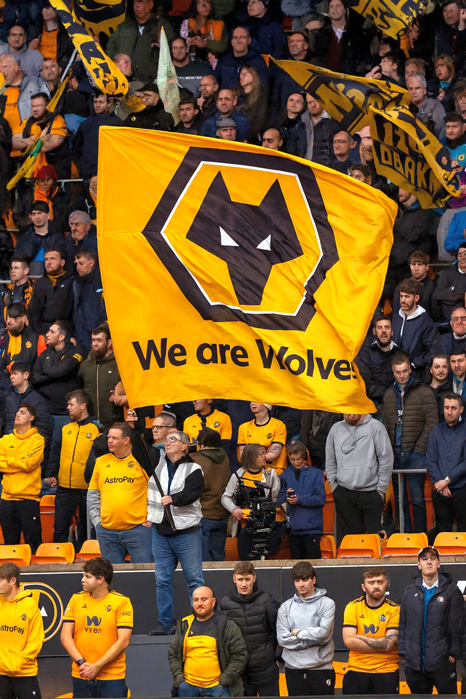 Une foule de fans de football dans un stade, avec un grand drapeau jaune des Wolverhampton Wanderers portant l'inscription 'We are Wolves' au premier plan.
