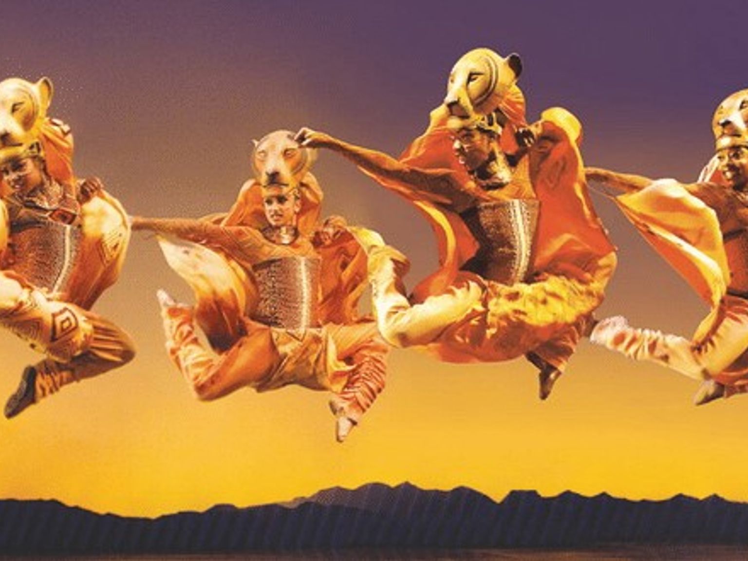 Fyra Lion King-artister i gyllene kostymer och lejonmasker hoppar mot en solnedgångshimmel.
