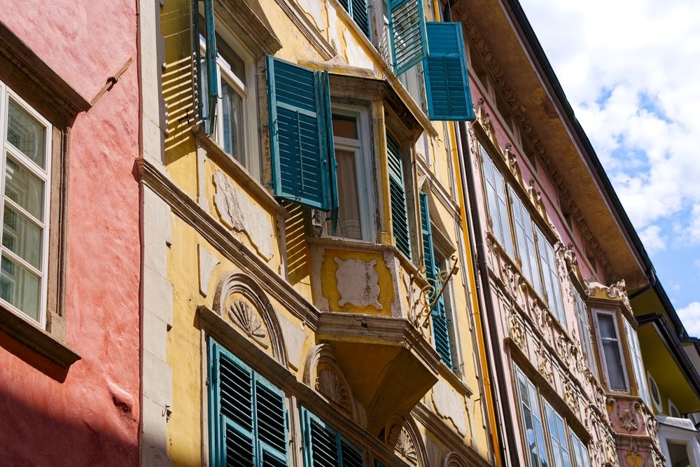 Prunkfulla gamla byggnader med flagnande gul och rosa färg, blå fönsterluckor och dekorativa fönster under en blå himmel. Bolzano, Italien