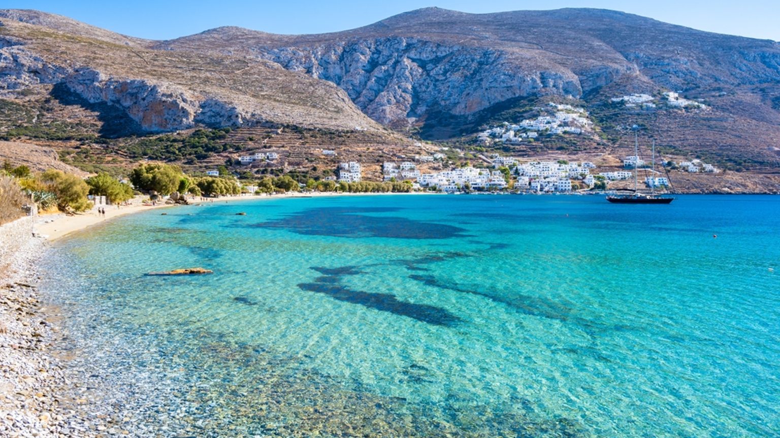Clear turquoise water in a bay with a pebble beach, a white village by the mountains, and a distant sailboat. Naxos, Greece