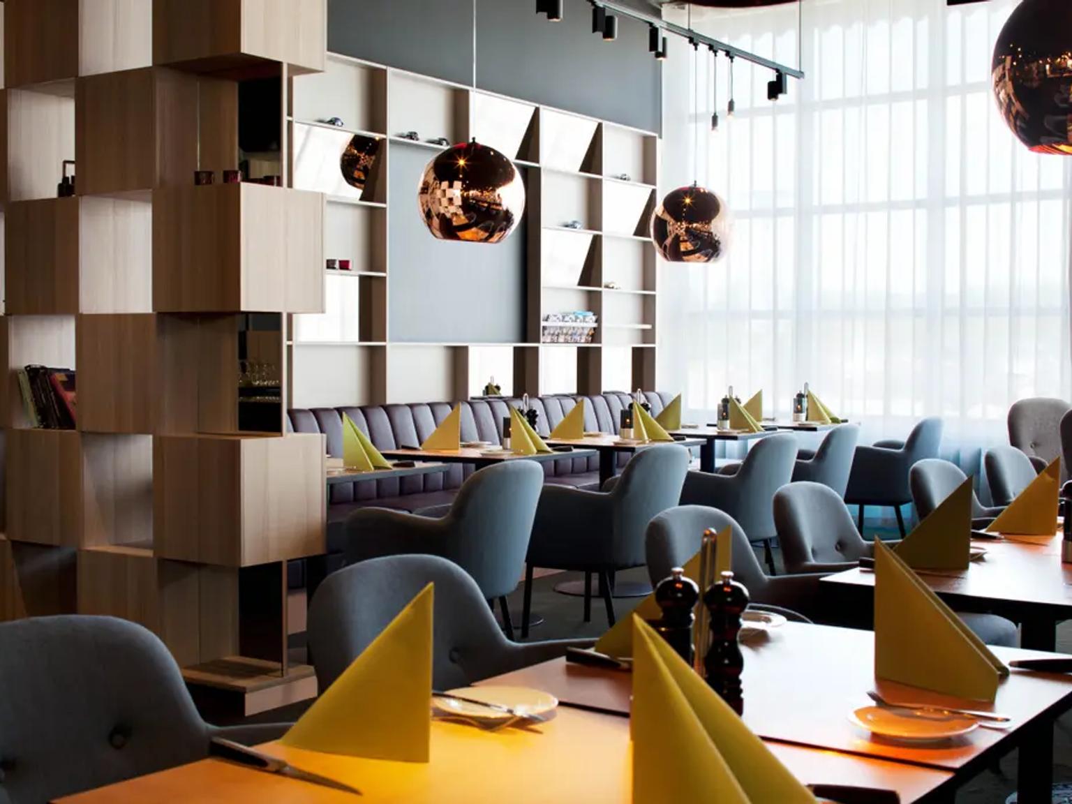Moderne restaurantinteriør med dekkede bord, grå stoler, lilla båser, en trevegg, kobberpendellamper og et stort vindu med lette gardiner. Scandic Kolding