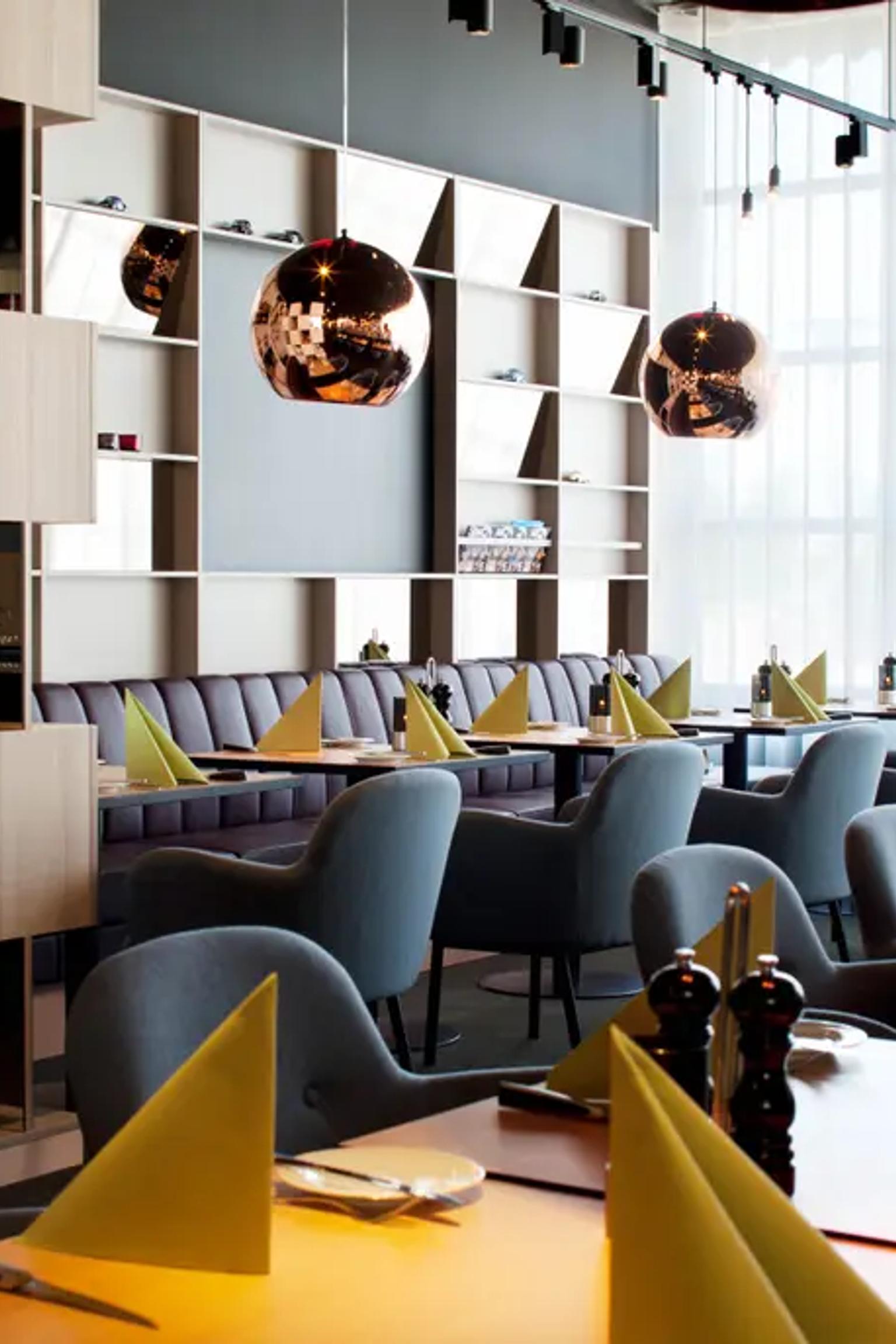 Moderne restaurantinteriør med dekkede bord, grå stoler, lilla båser, en trevegg, kobberpendellamper og et stort vindu med lette gardiner. Scandic Kolding