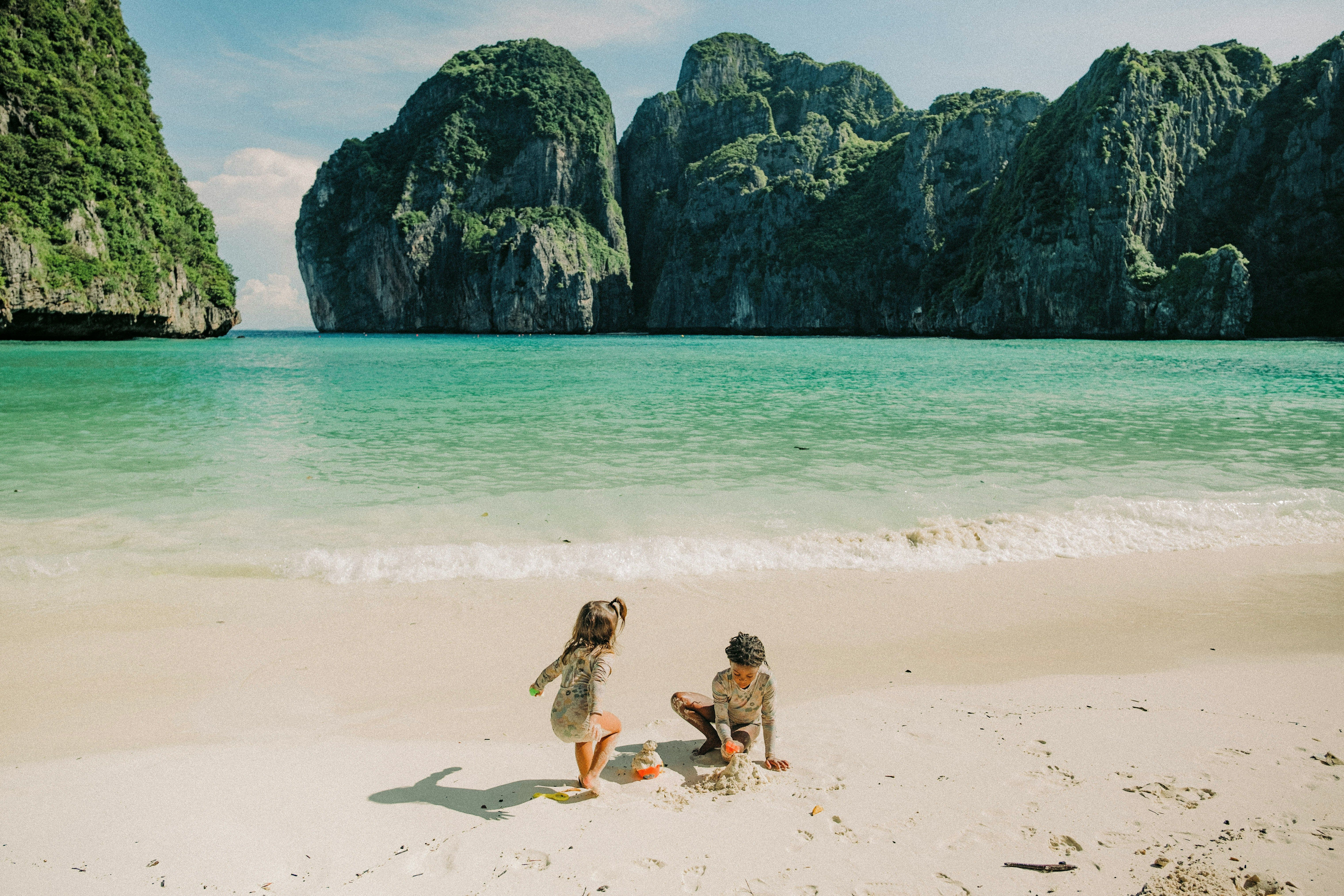 Två barn leker på en vit sandstrand med turkost vatten och höga gröna klippor i Thailand