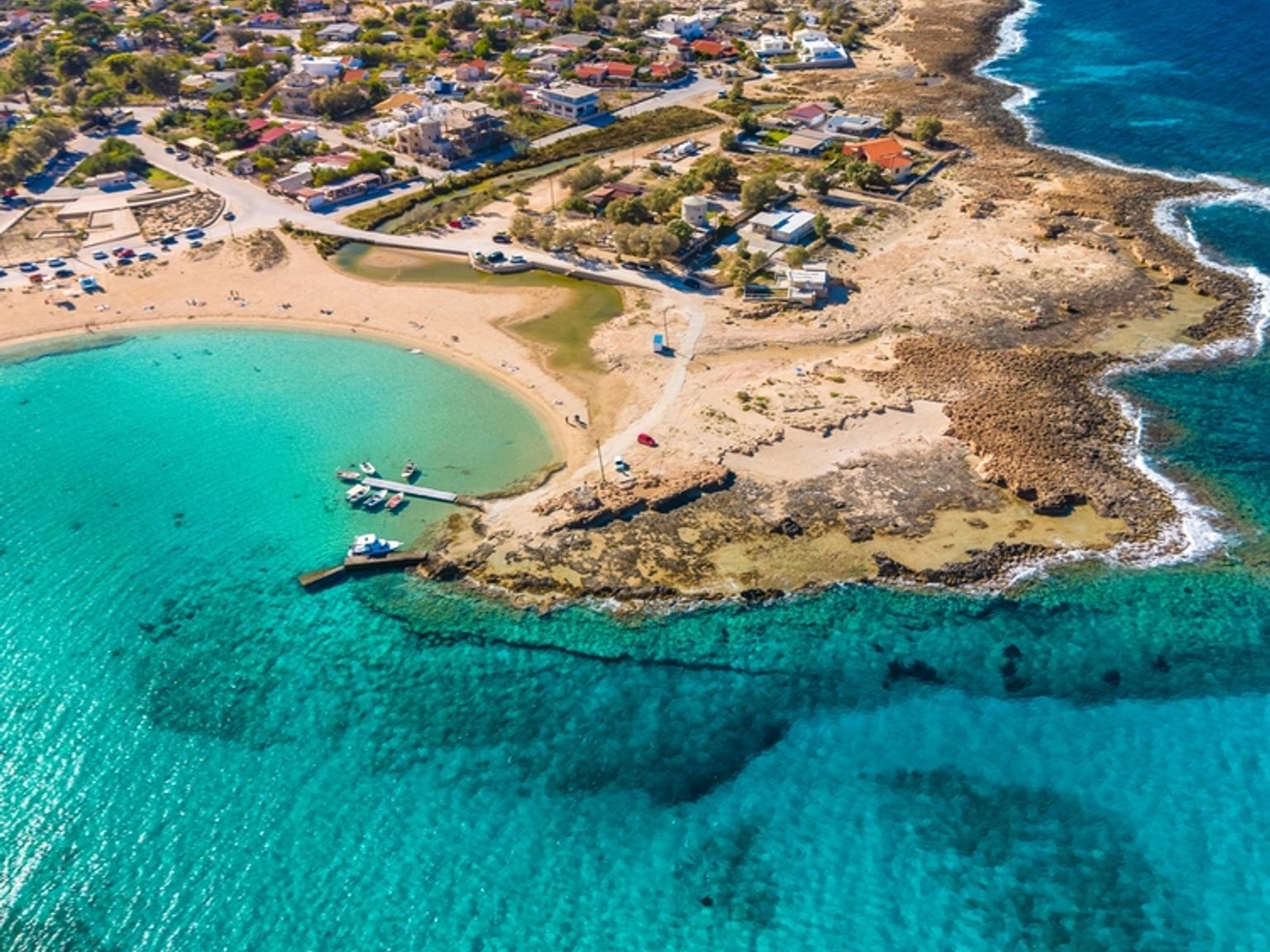 Luftfoto af en kystby med turkisblåt vand, sandstrande, klippefyldte kyster og både, der ligger fortøjet i en bugt.