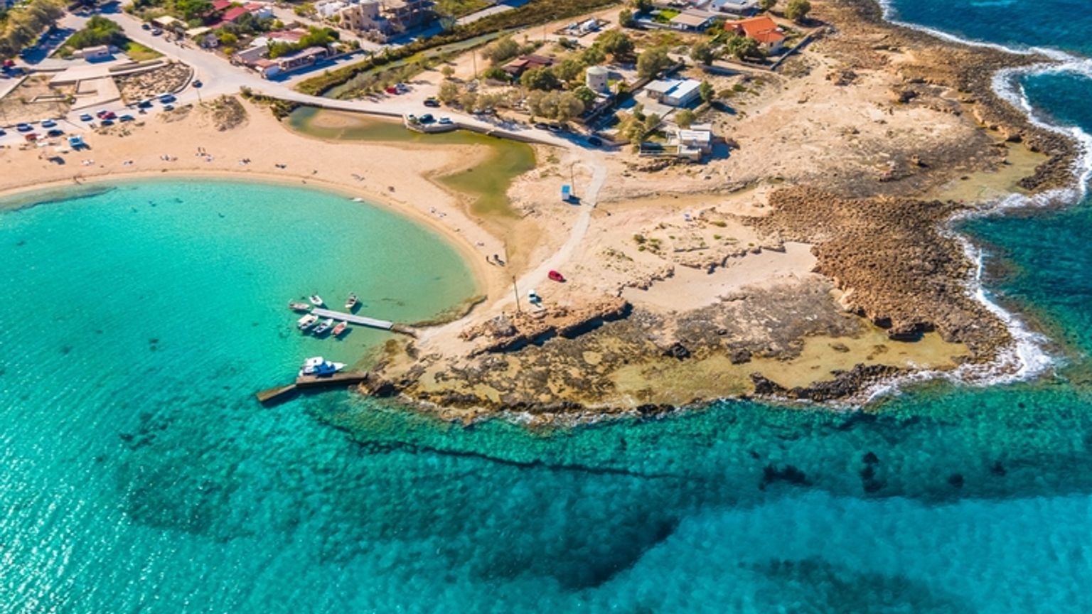 Luftfoto af en kystby med turkisblåt vand, sandstrande, klippefyldte kyster og både, der ligger fortøjet i en bugt.