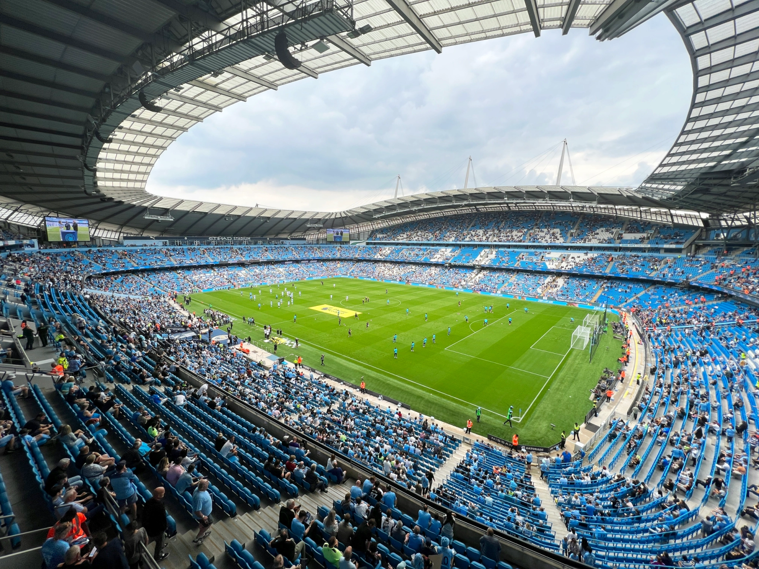 Match de football au stade Etihad, avec des joueurs sur le terrain et des fans dans les sièges bleus.