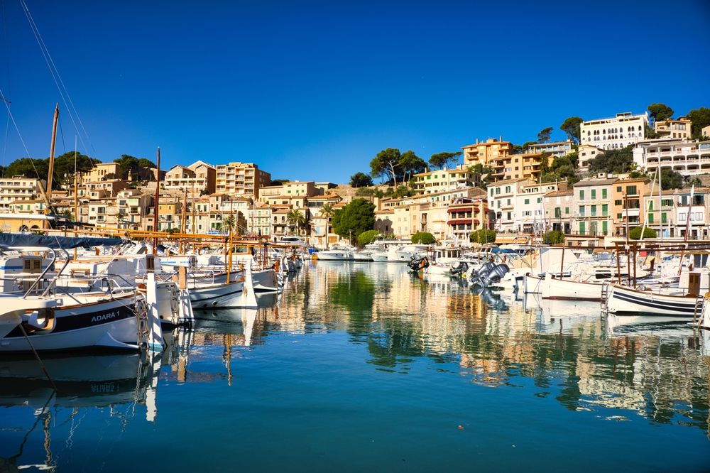En livlig havn med både og en farverig by på en bjergskråning under en blå himmel. Sóller, Mallorca