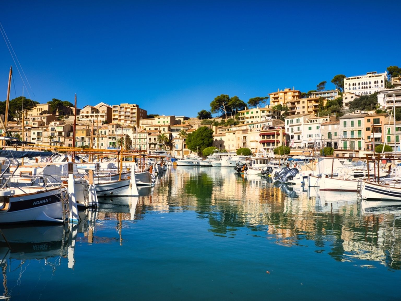 En livlig hamn med båtar och en färgstark stad på en sluttning under en blå himmel. Sóller, Mallorca