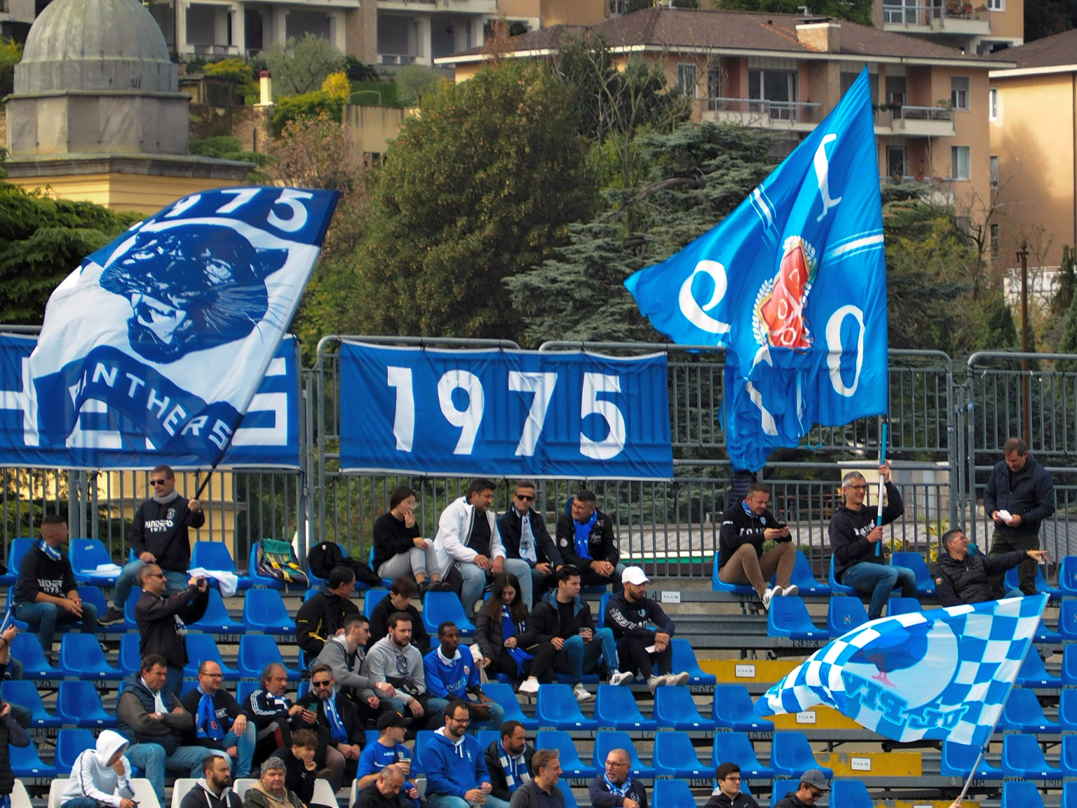 A stadium section with fans holding blue and white flags and banners displaying '1975' and a panther, overlooked by apartment buildings.