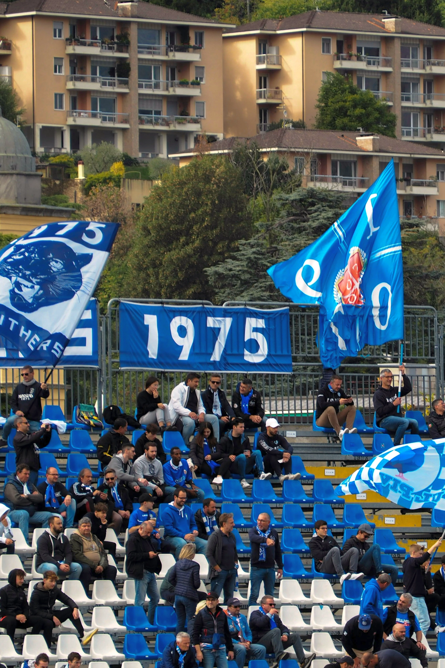 A stadium section with fans holding blue and white flags and banners displaying '1975' and a panther, overlooked by apartment buildings.