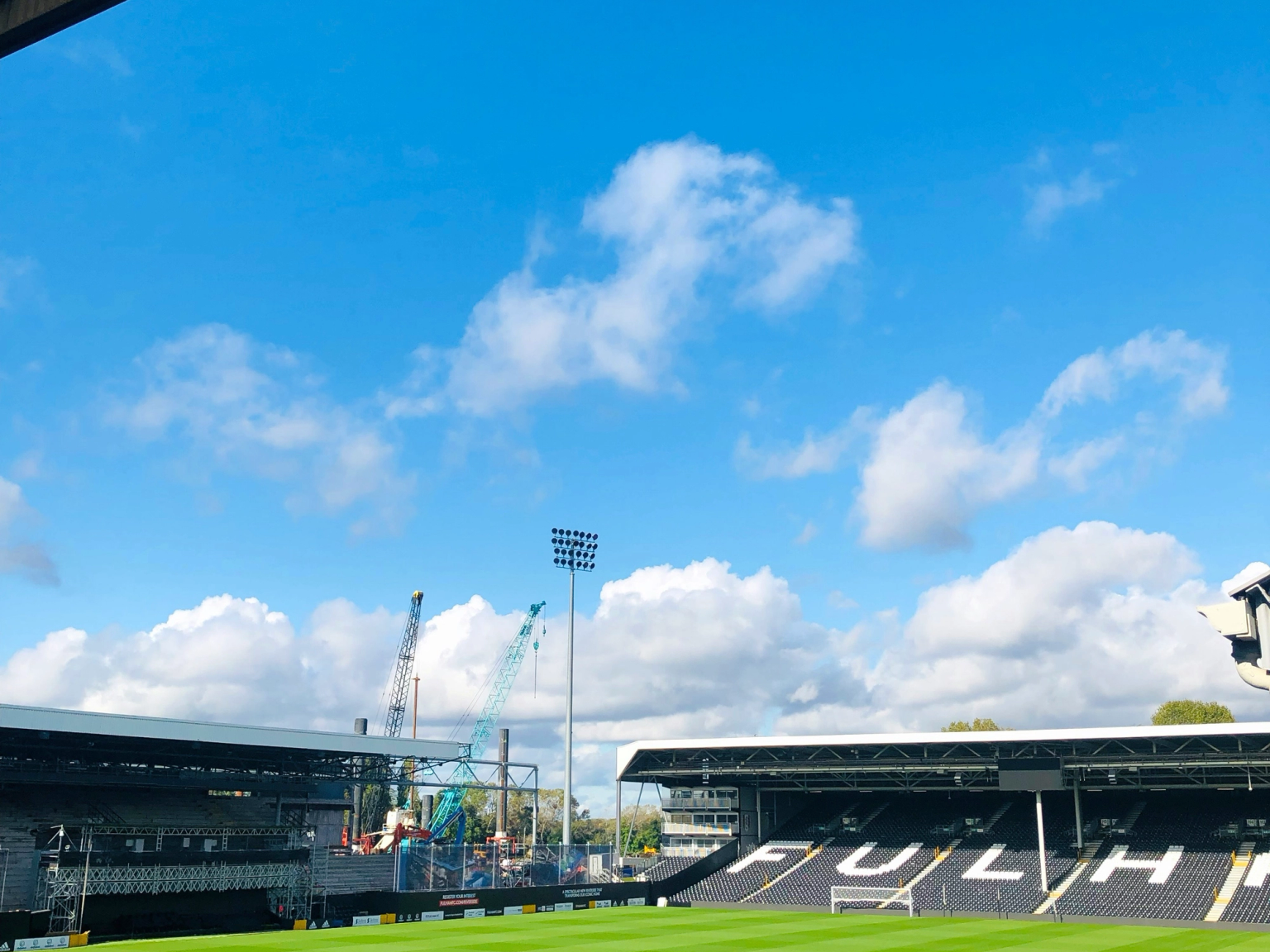 Een voetbalstadion met een groen veld, lege tribunes met de tekst "FULHAM" en bouwkranen onder een blauwe, bewolkte hemel.