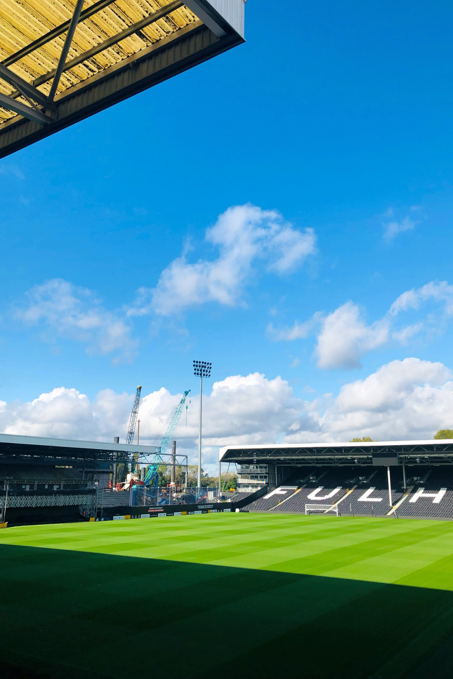 Een voetbalstadion met een groen veld, lege tribunes met de tekst "FULHAM" en bouwkranen onder een blauwe, bewolkte hemel.