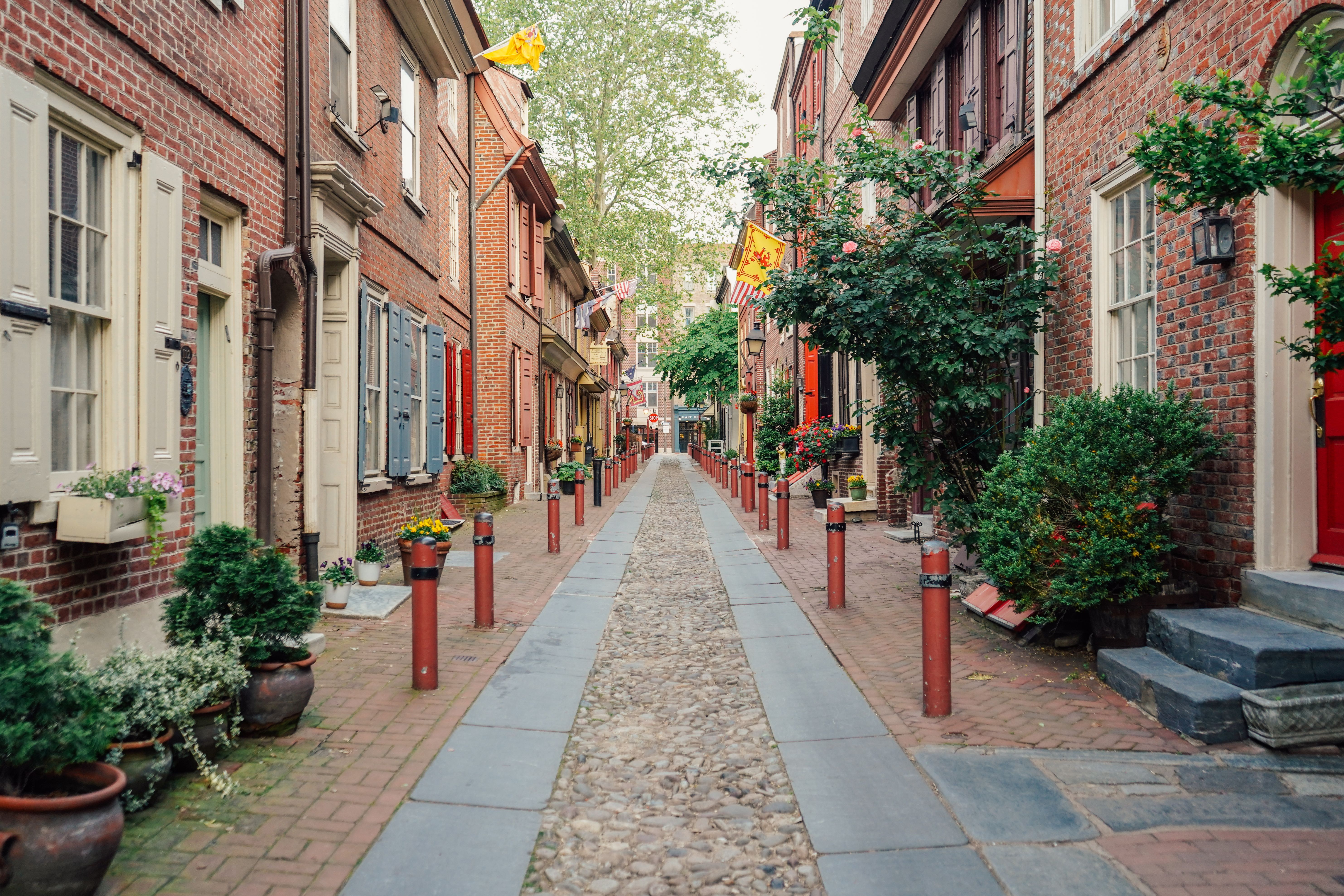 En smal brostensgyde flankeret af historiske rækkehuse i mursten med farverige skodder og potteplanter. Philadelphia