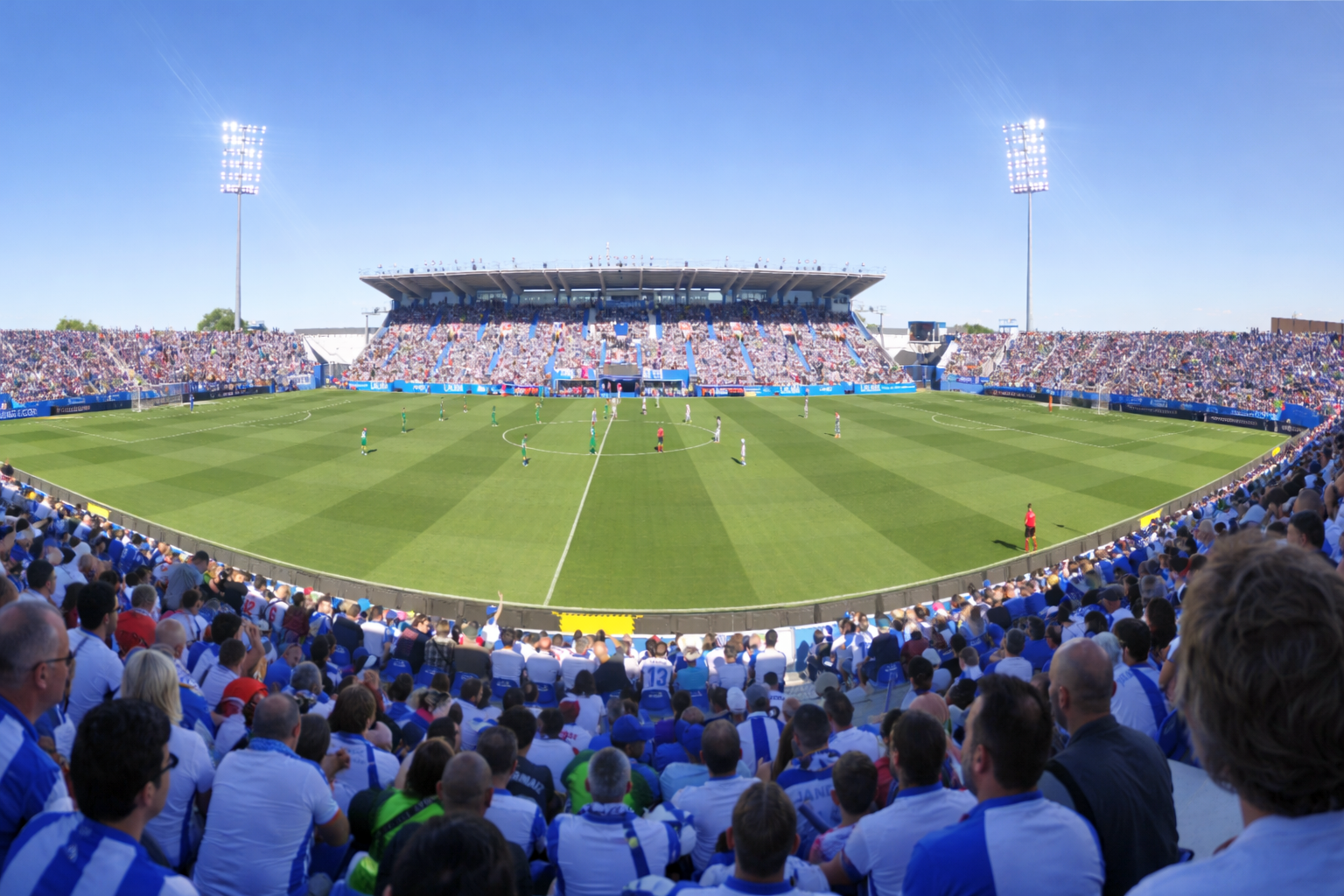 Estádio de futebol lotado durante uma partida diurna, visto das arquibancadas.