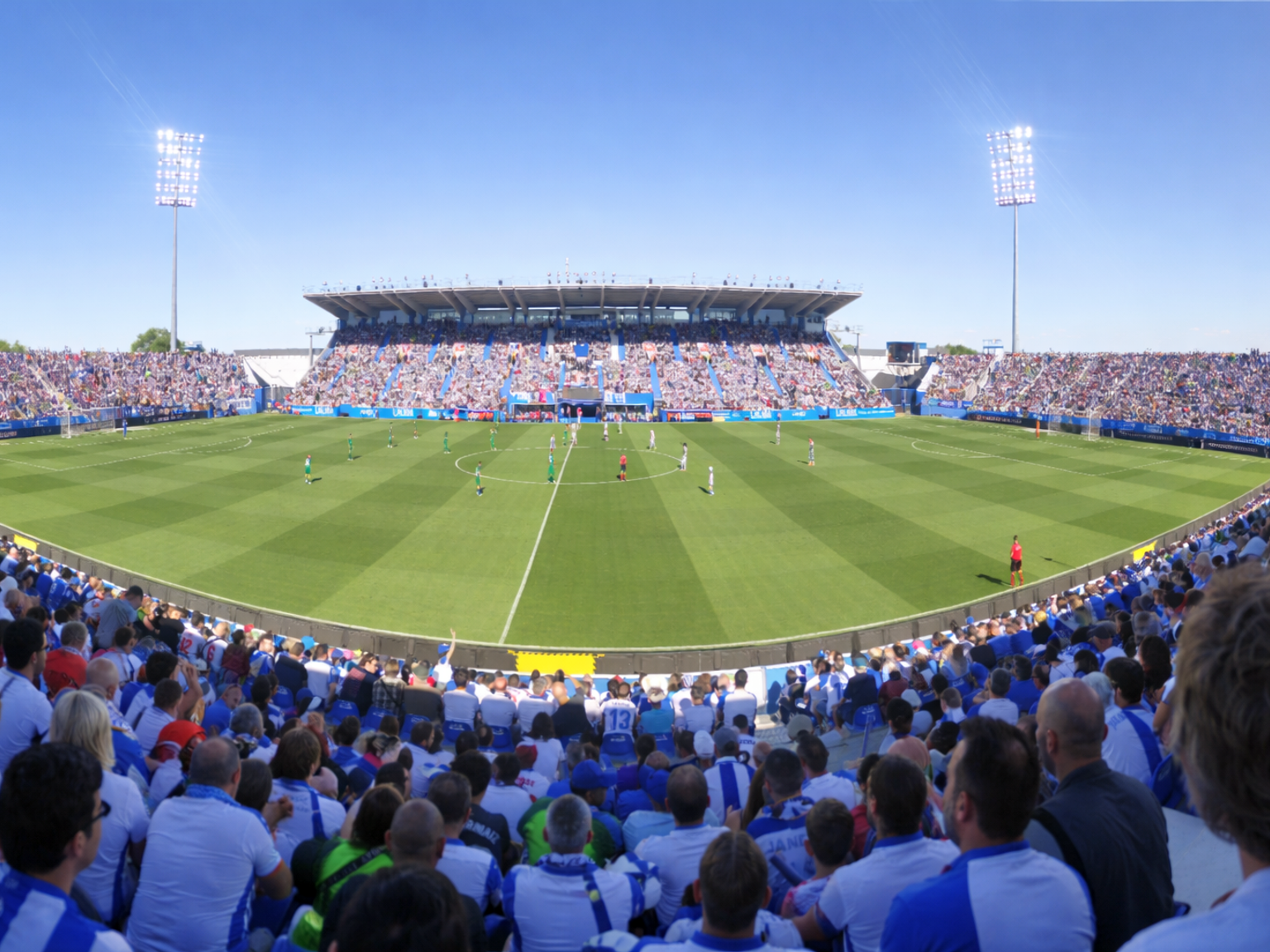 Estádio de futebol lotado durante uma partida diurna, visto das arquibancadas.